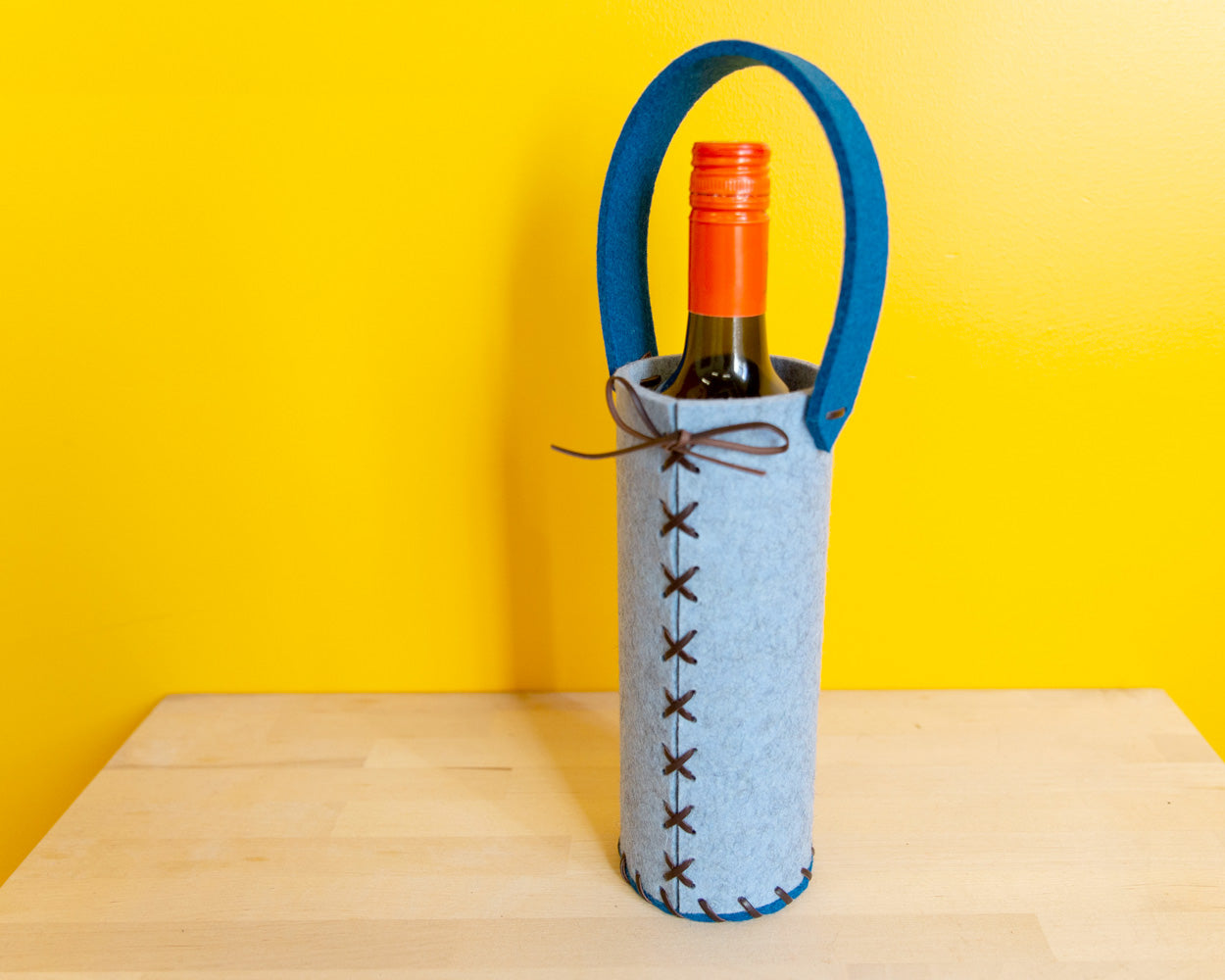 Side view of a light and dark blue felt wine tote with leather lacing and a dark wine bottle inside, against a yellow wall.