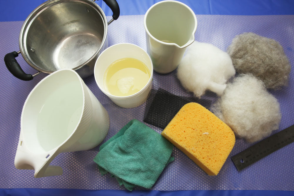 A top down view of wet felting supplies on a table. From left to right: a milk bag holder filled with water, a small pot, a container of liquid soap, a green cloth, a tall plastic container, black screen mesh, a large yellow sponge, white, off-white, and beige carded wool, a metal ruler.