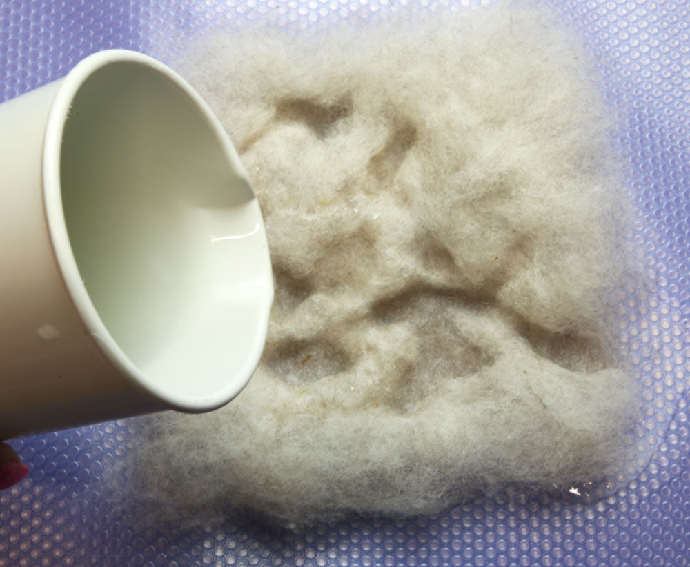 A top down view of a white plastic container gently pouring water over a square of off-white carded wool.