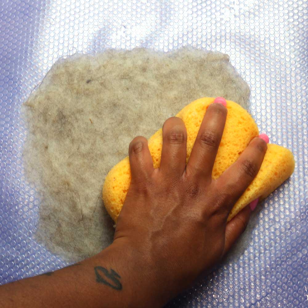 A hand holding a yellow sponge pressing down on a square of wet beige wool.