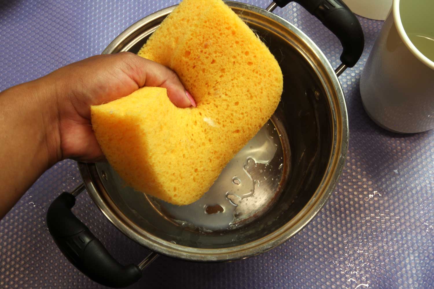A hand squeezing a yellow sponge over a pot to empty water from the sponge.