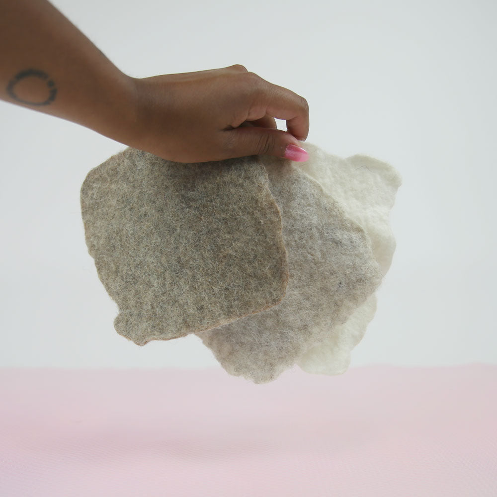 A hand holding three wet felted squares of varying beige tones above a pink table