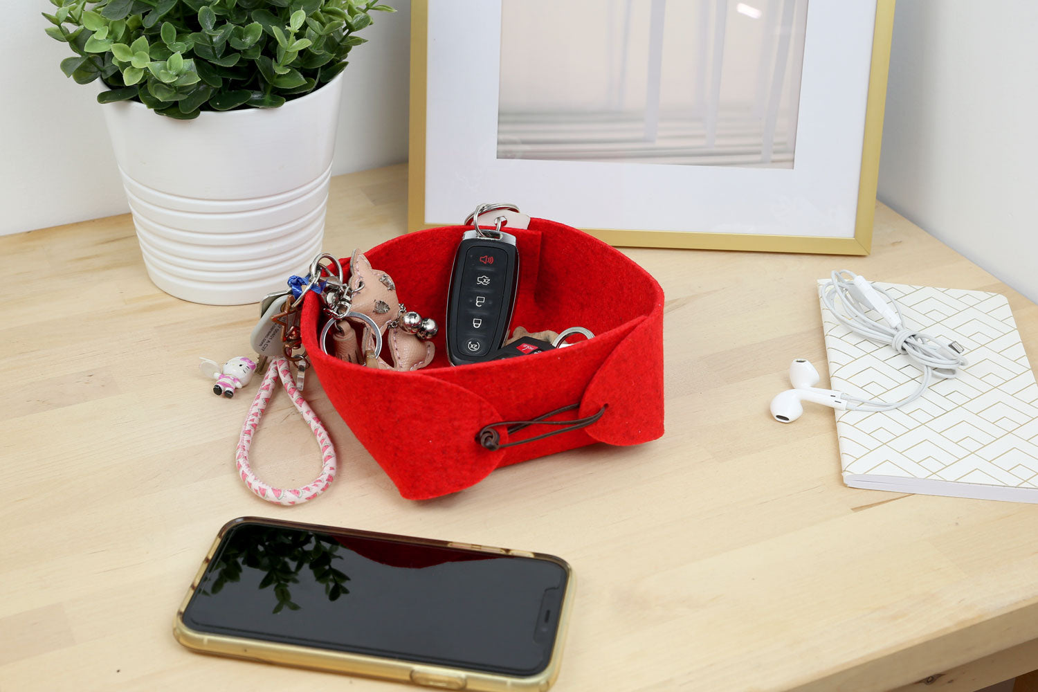 A red felt box with leather cord sitting on a wooden table. There are keys inside the box. A phone, notebook, and a pair of earbuds are next to the box.