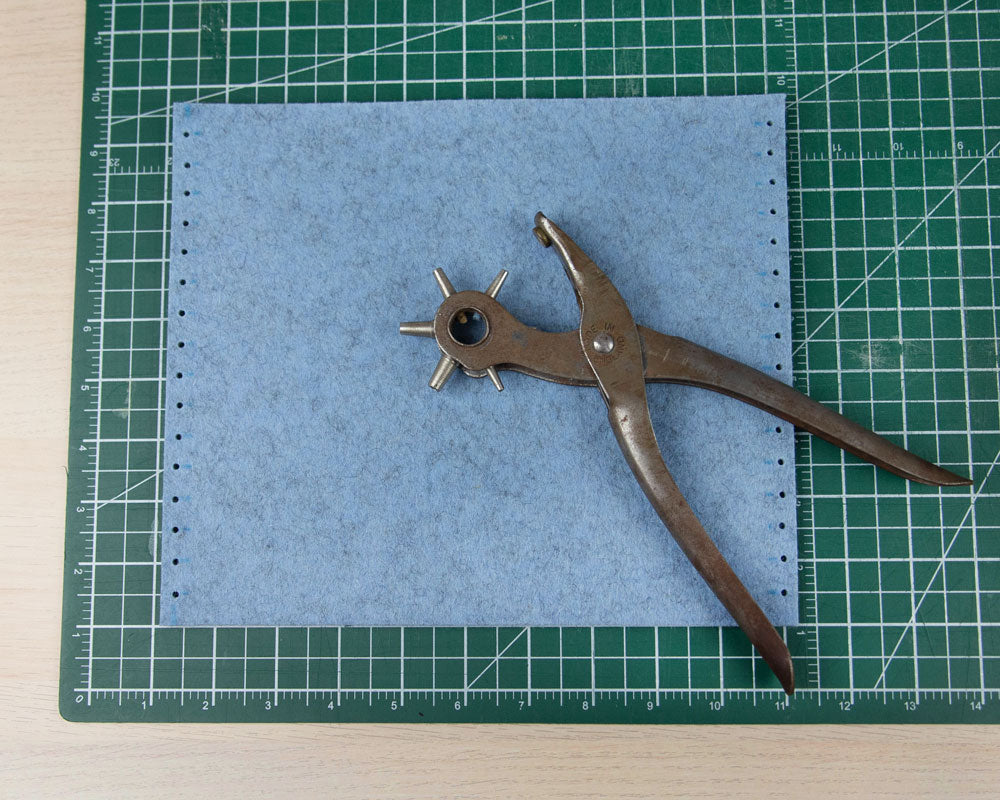 An overhead shot of a rectangle of blue felt with several small holes on the left and right sides sits on a green cutting mat. A leather hole punch lays on the blue felt. 