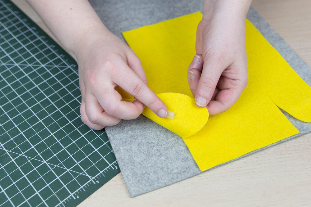 A pair of hands are placing masking tape to a felt box template to keep it in place on a piece of thick grey felt