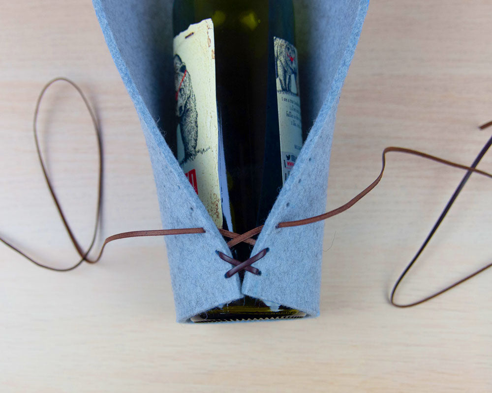 A dark wine bottle on its side with blue felt wrapping around it. There are hole on the edges of the felt. Leather cord is laced in the first three holes at the bottom, keeping the edges together.