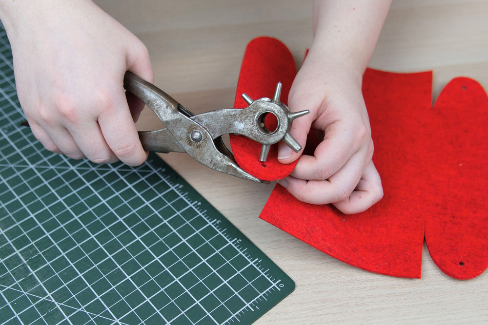 A pair of hands holds a leather hole punch and a piece of red felt. The hole punch has just made a hole in the felt.