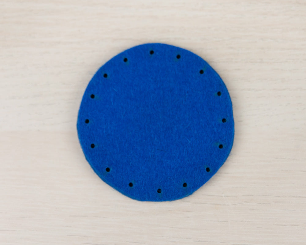 A circle of dark blue felt on a light wood table. There are small holes punched around the edge of the circle.