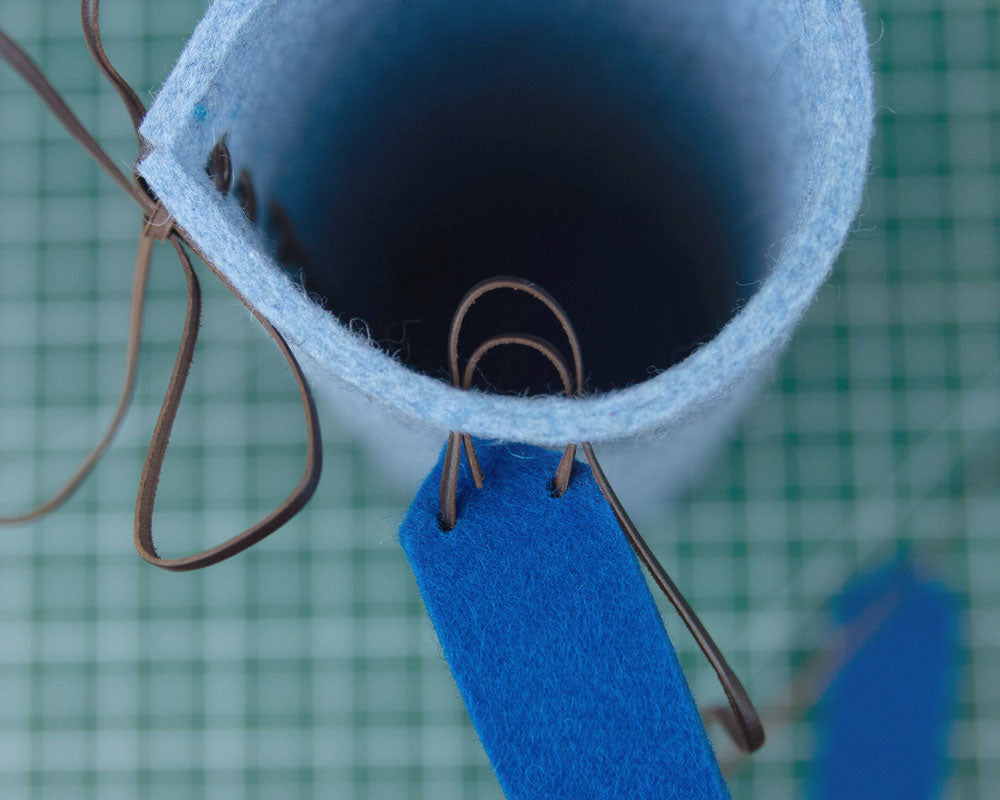 A top down shot of a cylinder of light blue felt. A strip of dark blue felt is loosely laced to the side of the cylinder with leather cord.