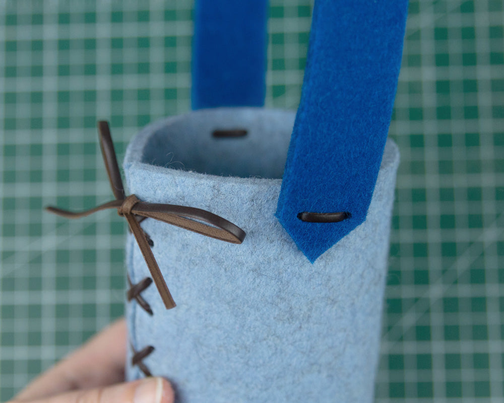 A side view of a light blue felt cylinder with a dark blue felt handle. leather cord laces up the cylinder and attaches the handle to the sides of the cylinder.