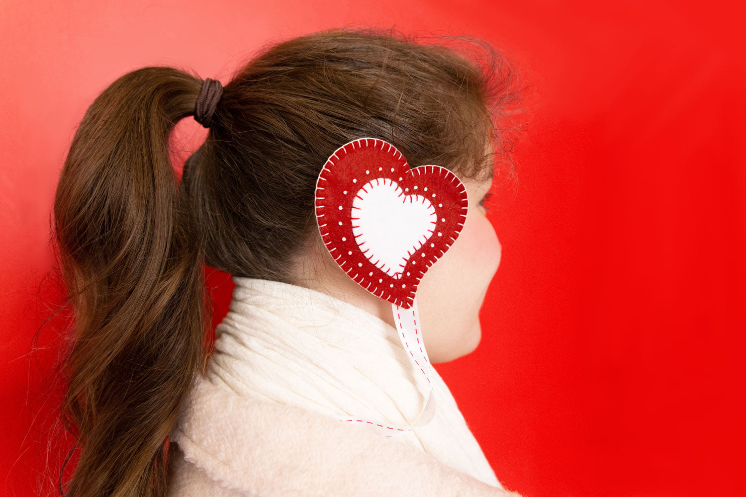 A profile shot of a person with a long, blonde, high pony-tail in a pink coat and white scarf on a red background. They are wearing a red and white heart shaped earmuff.