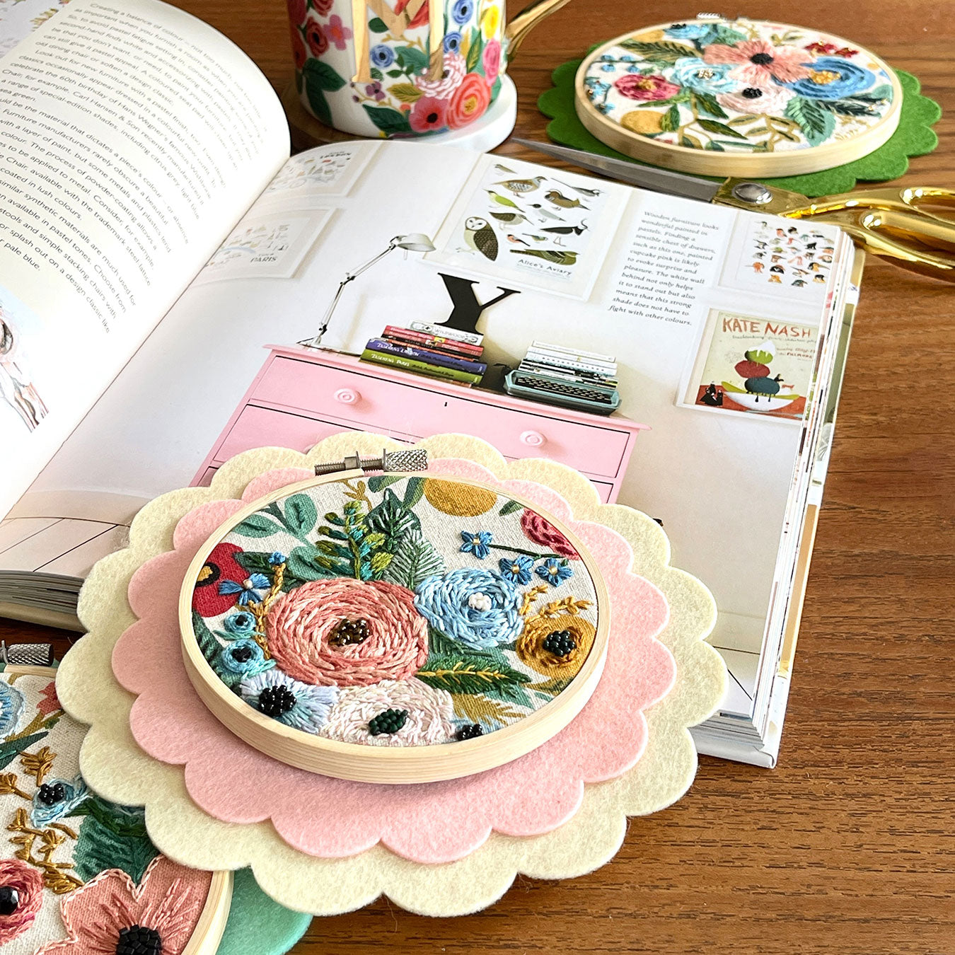 An embroidery hoop with a scalloped felt backing sits on an open book on a wooden table.