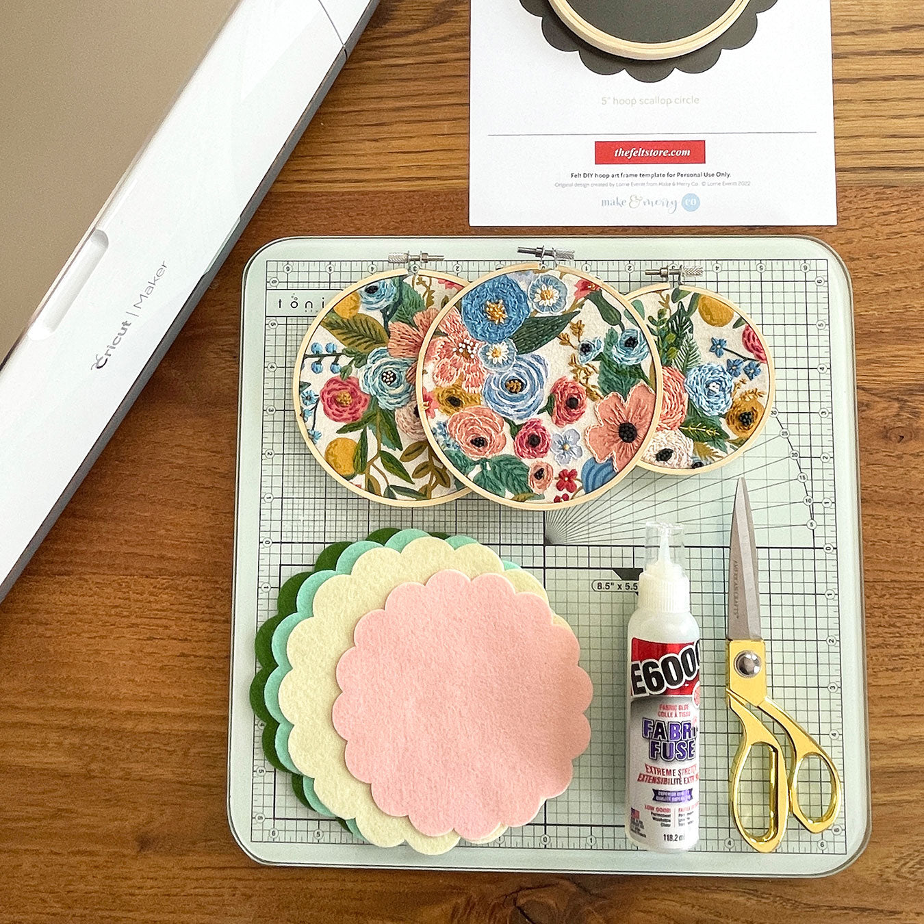 Three embroidery hoops, scalloped felt circles, scissors, and a bottle of E6000 fabric fuse lay on a small cutting mat.
