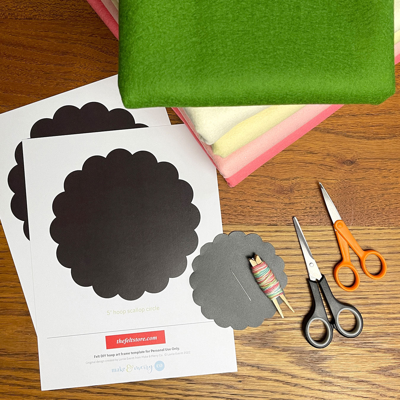 Printer paper templates with scalloped circles and two pairs of scissors lay on a wooden table. A pile of felt material is visible from the corner.