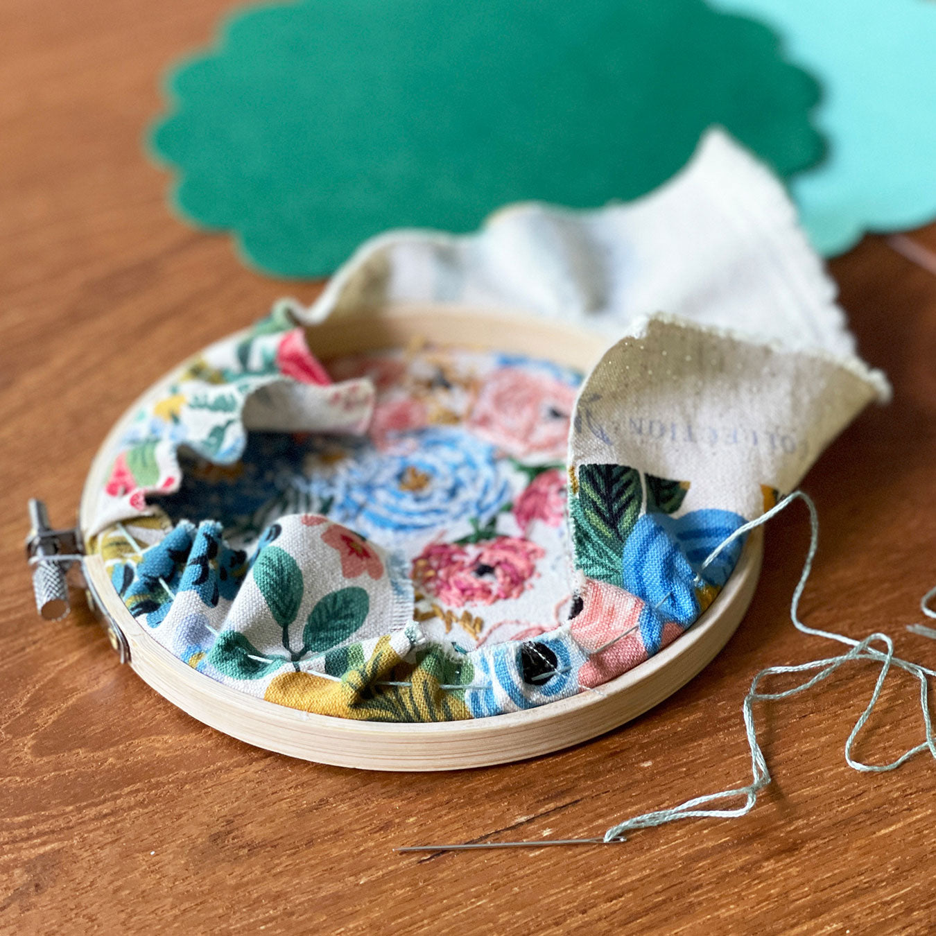 An embroidery hoop with fabric is laying face down on a wooden table. A sewing needle with thread has sewn and gathered a circle around the edge of the material at the back of the hoop.