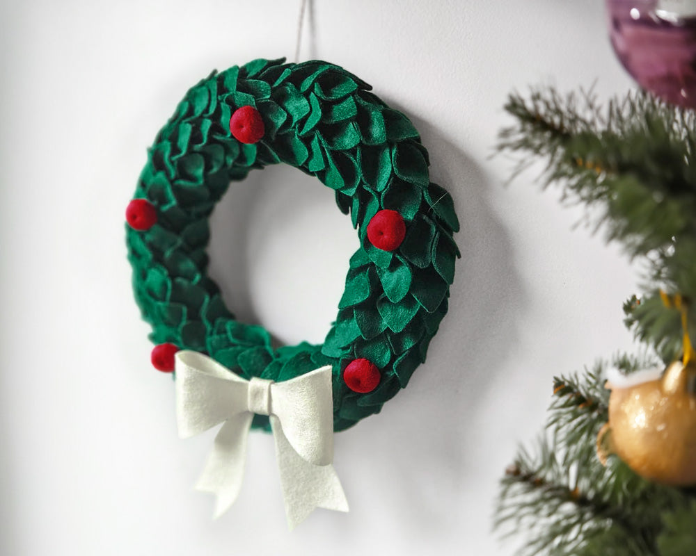 Completed DIY Felt Christmas Wreath hanging on a plain wall with a Christmas tree in the foreground