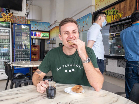 Simon is eating some Kaya toast while holding a cup of Kopi-O at a coffee shop