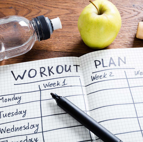 Notebook with workout plan schedule drawn in surrounded by water bottle and apple