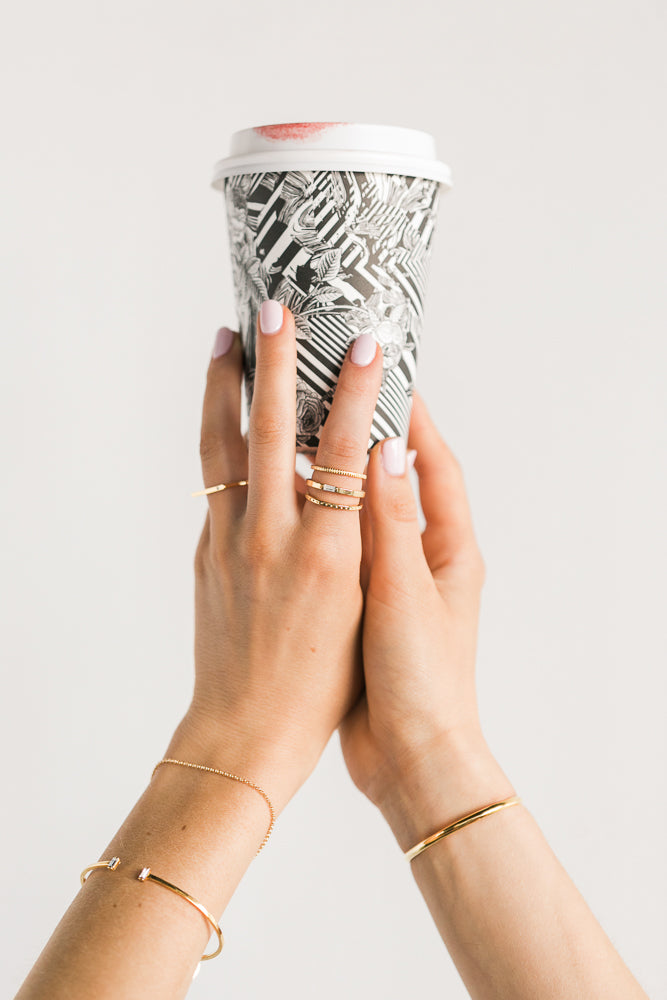 Hand holding a stylish patterned takeaway coffee cup adorned with Katie Dean Jewelry stacked gold rings, showcasing minimalist jewelry design.