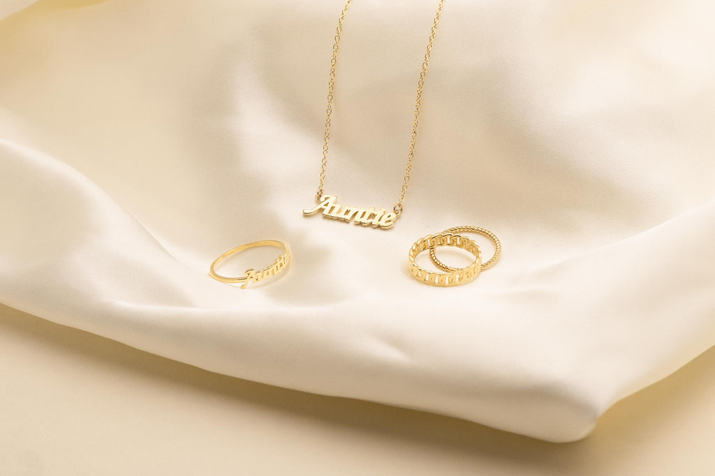 Picture of a dainty gold Auntie Necklace laying on satin alongside gold stacking rings, made in America by Katie Dean Jewelry 