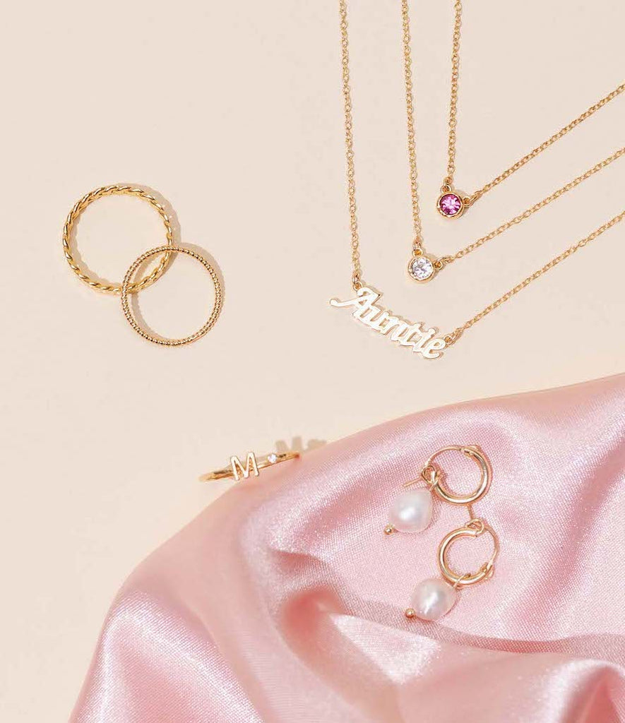 Dainty gold Birthstone Necklaces layered with the Auntie Necklace on a tan background with dainty stacking rings styled around the necklaces and a pair of gold pearl hoops from the Katie Dean Jewelry collection.