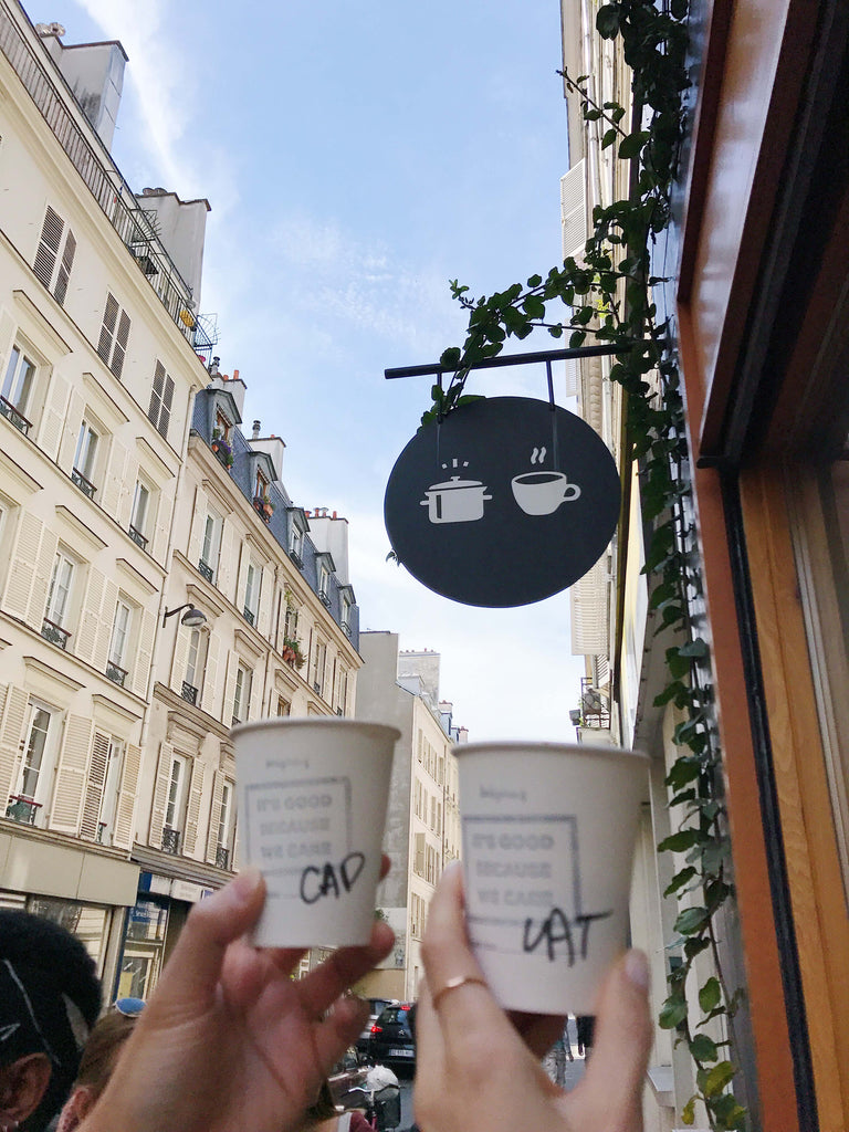 Cheerful toast with coffee cups under a quirky café sign in Paris, embodying the joy of shared moments and good beverages while showcasing a dainty gold band ring by Katie Dean Jewelry.