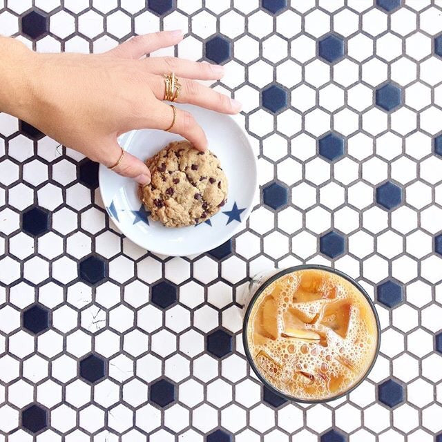 Woman's hand with delicate rings over a plate with a cookie and an iced latte on a hexagonal tiled table, blending refreshment with fashion.