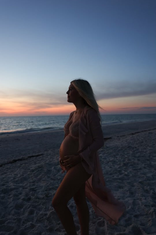 Katie Dean beach maternity photo shoot, six and a half months pregnant holding her baby bump with a sunset and the ocean in the background.