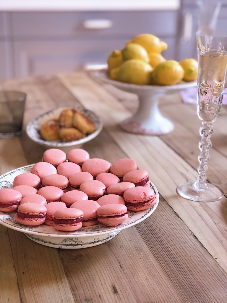 Pink French Macarons on a vintage plate on a wooden table fresh from the oven with a dish of lemons in the background and a glass of champagne next to it.