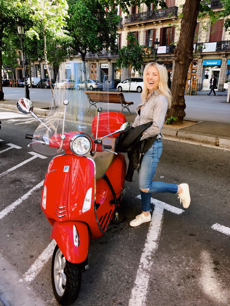 Katie standing next to a red moped motorcycle smiling wearing jeans, white shoes and a striped button down shirt, in Barcelona, Spain.