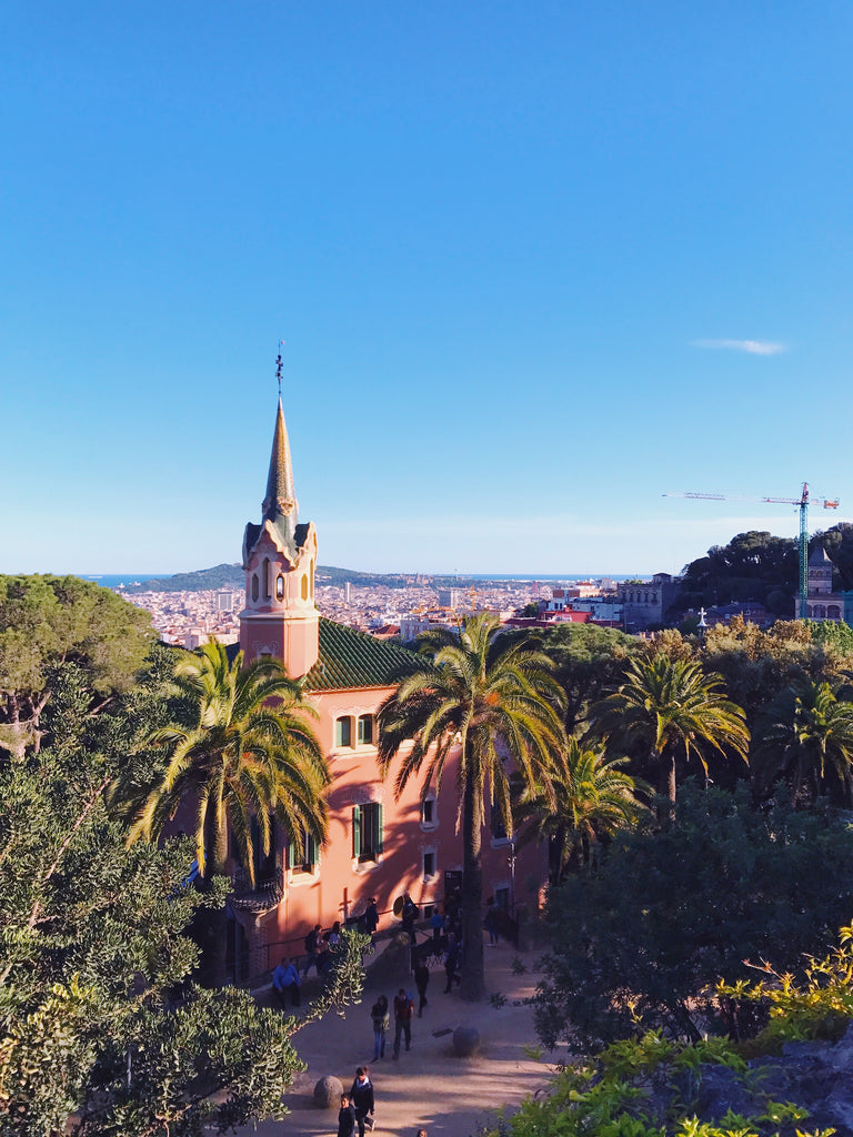 View of Barcelona from Park Guell. Showing a blue clear sky, palm, trees and a stunning pink building with a pointed steeple.