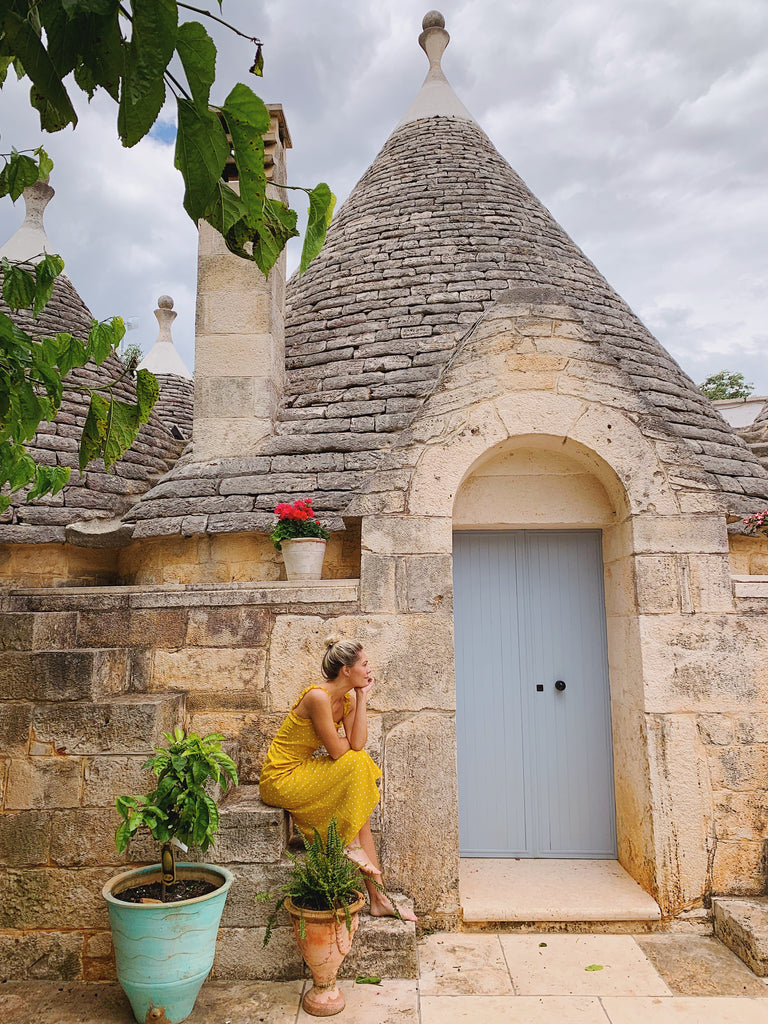 Katie Dean sitting on the steps of Trullo Sant'Angelo in a yellow dress 