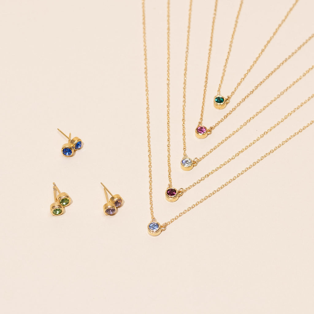 Assorted handmade gold birthstone necklaces and matching stud earrings made by Katie Dean Jewelry on a beige background.