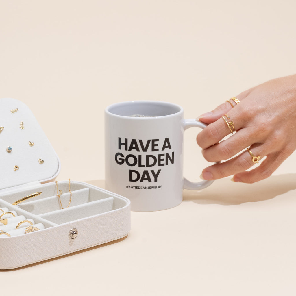 Elegant hand holding a 'Have a Golden Day' coffee mug next to a jewelry box, representing Katie Dean's unique dainty stacking ring collection.