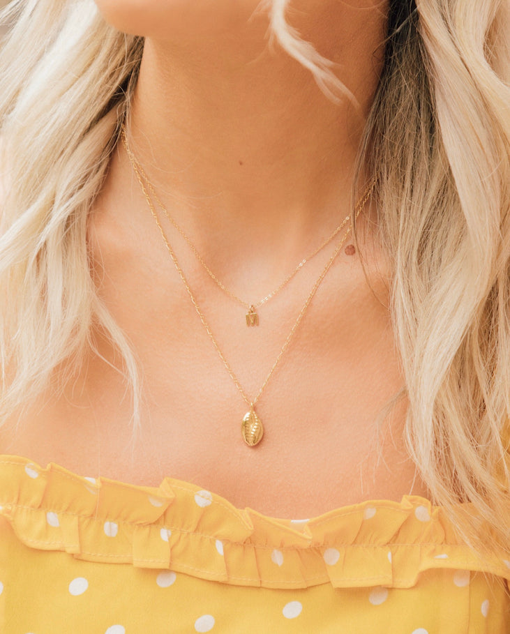 Layered gold necklaces featuring an 'M' initial pendant and a puka shell on a woman wearing a yellow ruffled top.