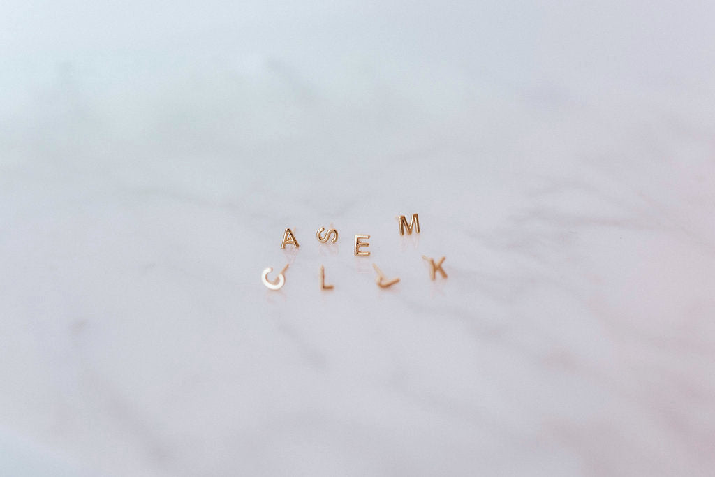 Gold initial stud earrings by Katie Dean Jewelry arranged on a marble surface, featuring letters A, S, E, M, L, and K.
