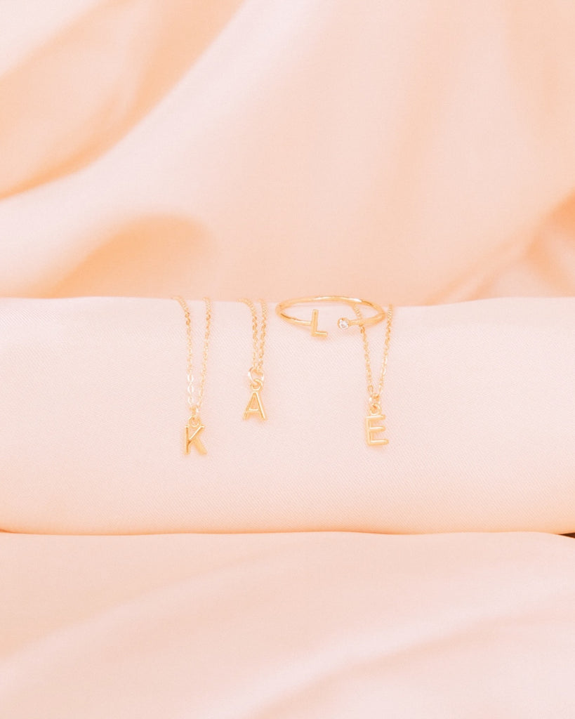 Katie Dean initial necklaces spelling 'KAE' and an initial 'L' ring displayed on pink satin fabric.