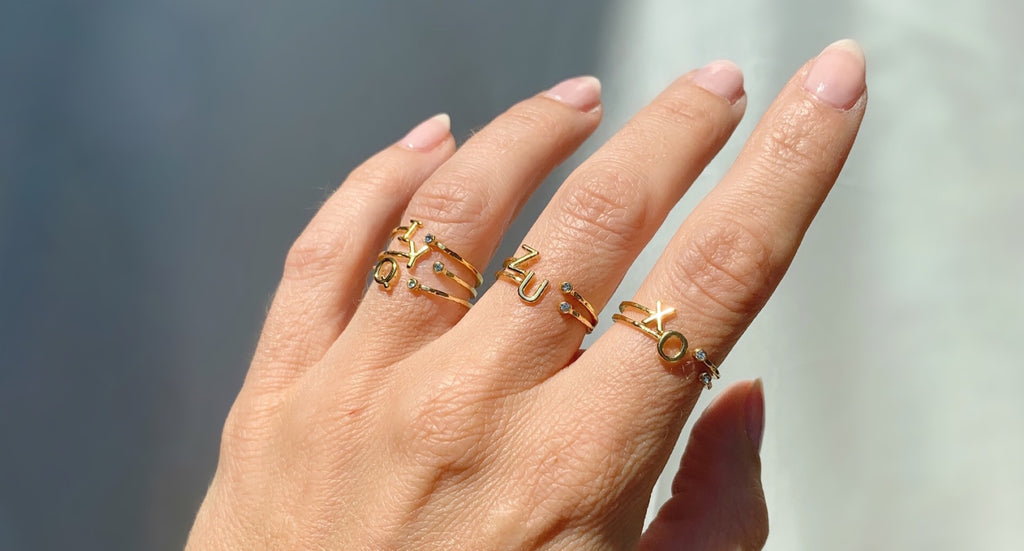 Hand displaying a selection of Katie Dean Jewelry's dainty letter rings with initials I, O, Q, U, X, Y, and Z in gold with white Swarovski crystals..