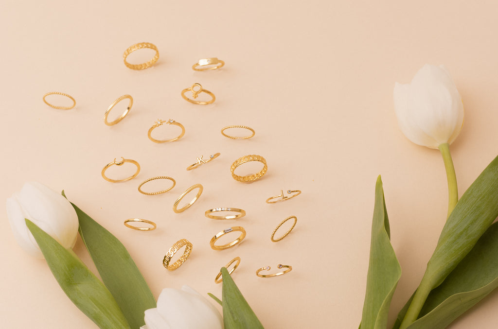 Assorted 14K gold-plated rings by brand Katie Dean Jewelry in various designs, including textured, smooth, and gem-set, elegantly displayed on a soft peach background with white tulips and lush green leaves.