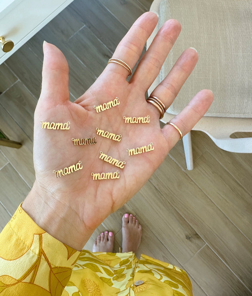 Gold Mama Charms on the palm of Katie's hand glowing from the natural light shining in. These dainty charms are made in America.