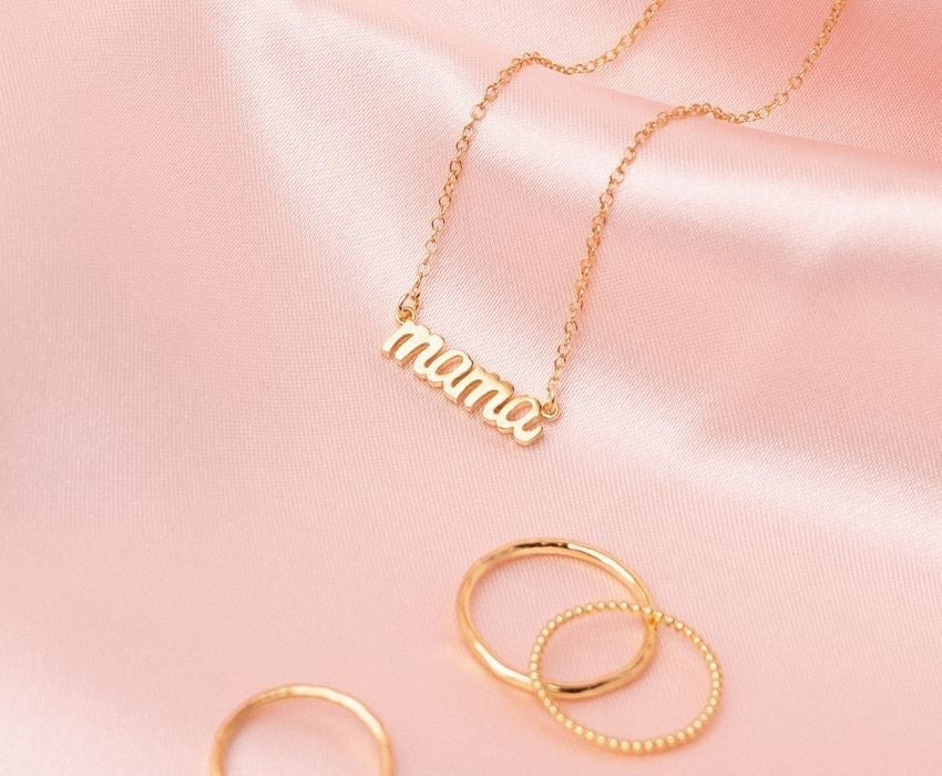 Gold necklace with the word 'Mama' crafted by Katie Dean Jewelry, as displayed on pink satin..