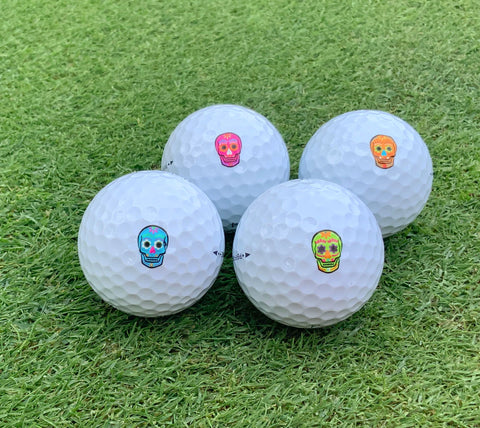 Four white golf balls with multi-colored skull stickers on grass.