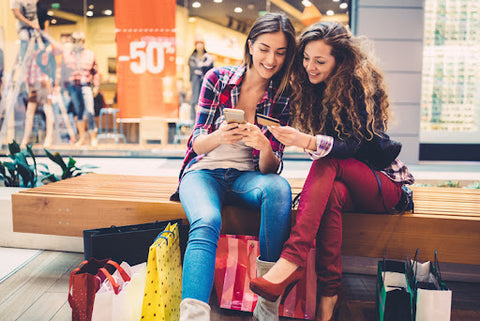 Two women at the mall sitting on a bench smiling at phones