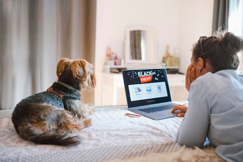 Woman lying in bed with her dog looking at Black Friday sales on computer