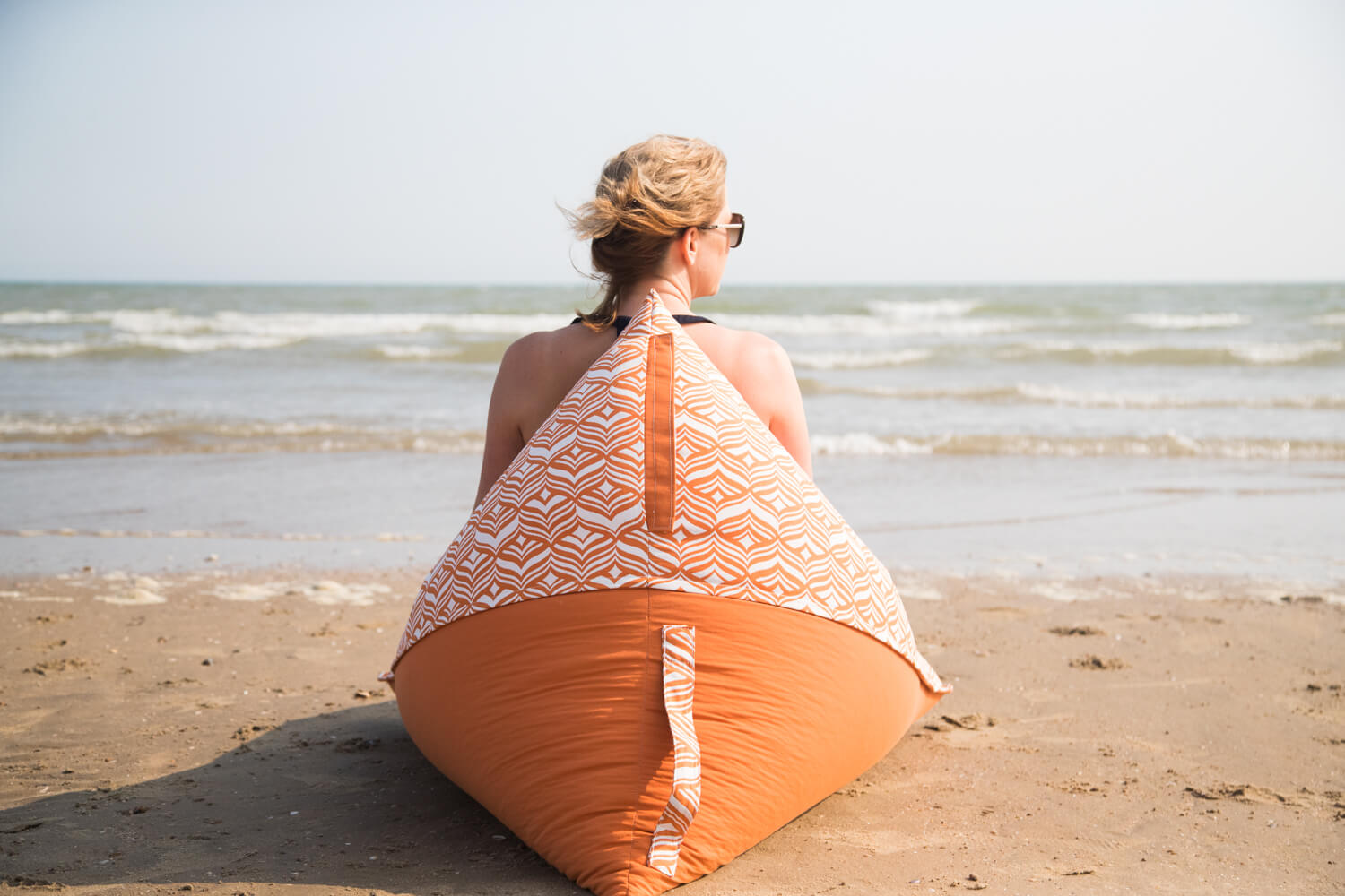 A woman sits on an orange beach lounger beanbag looking out at the waves