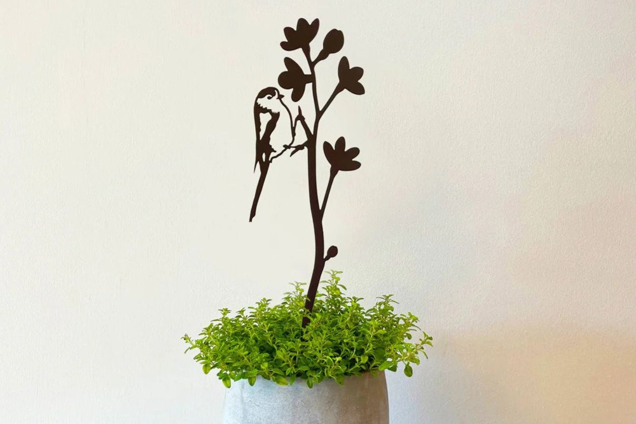 A metal silhoette of a long-tailed tit feeding on flower blossom has been put in an indoor pot filled with a plant with some light green leaves.