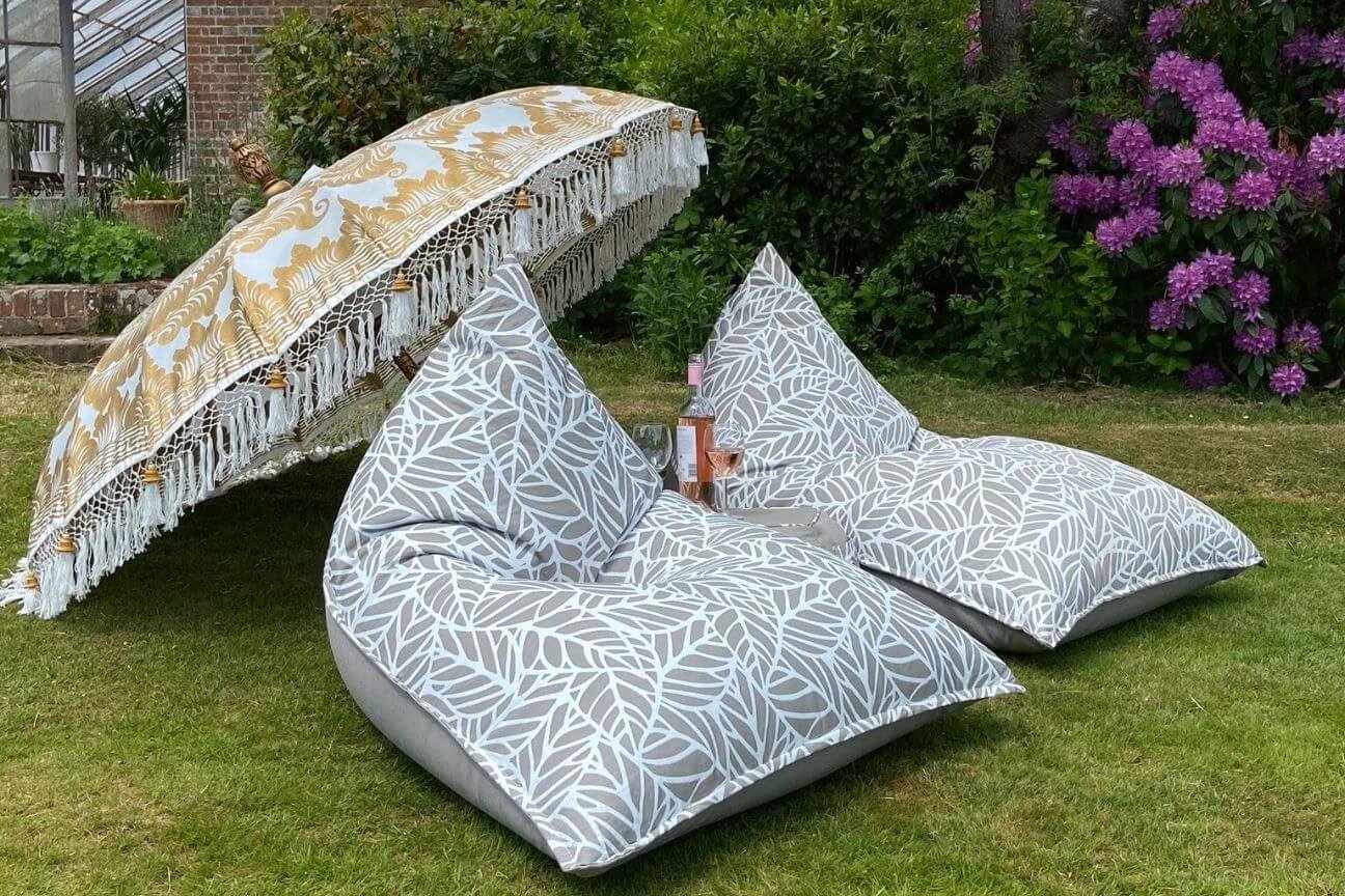 Two light-coloured grey palm-patterned bean bags placed on a lawn under a delicately patterened parasol
