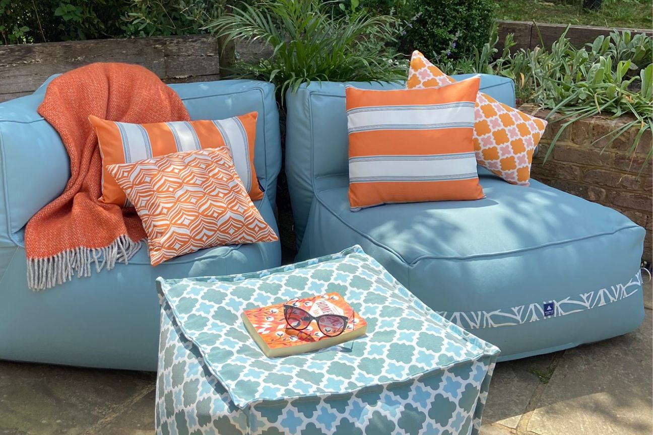 Two soft blue garden chairs with orange striped garden cushions in a patio corner. There is a blue geometric-patterned pouffe in the foreground with sunglasses and a book.