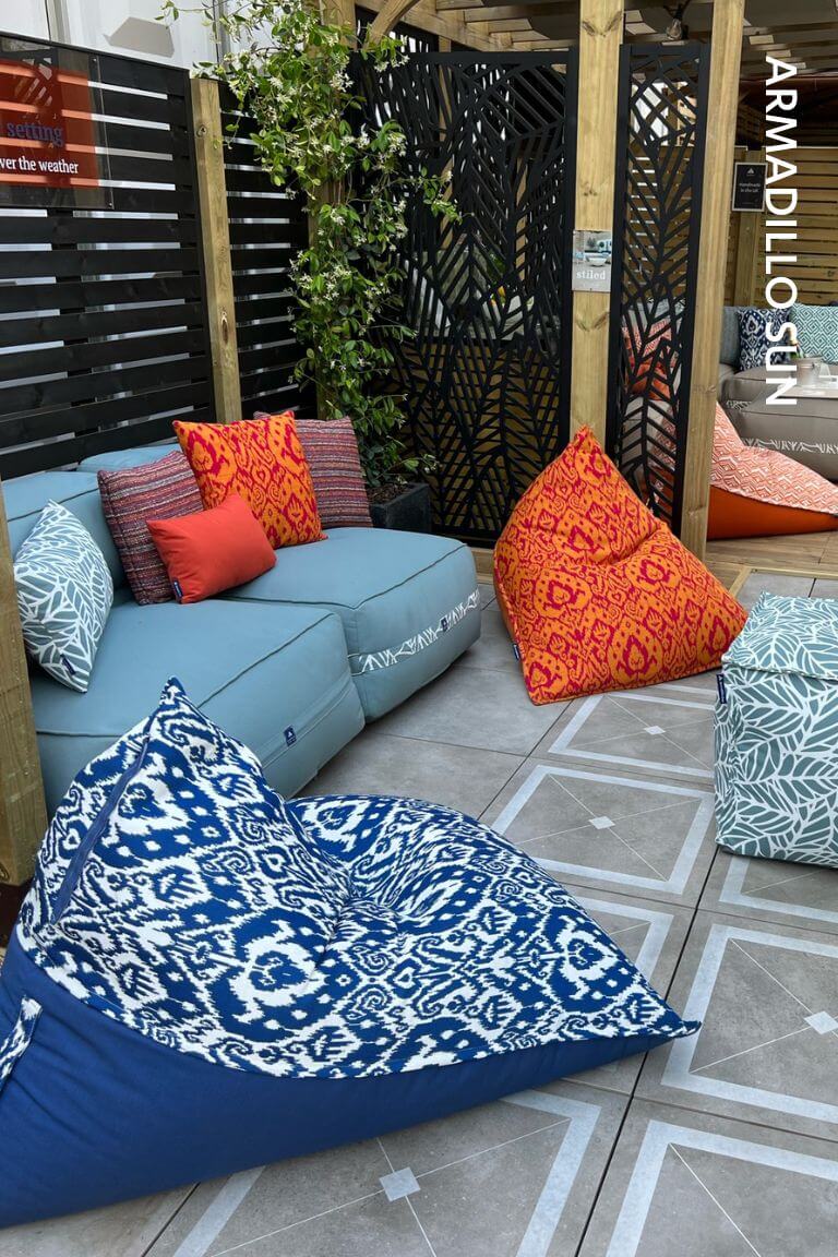 A photograph of a small tiled patio garden furnished with an ocean blue outdoor fabric sofa, two brightly coloured and patterned garden bean bag chairs, and a waterproof pouffe table. Text down the side of the image reads Armadillo Sun