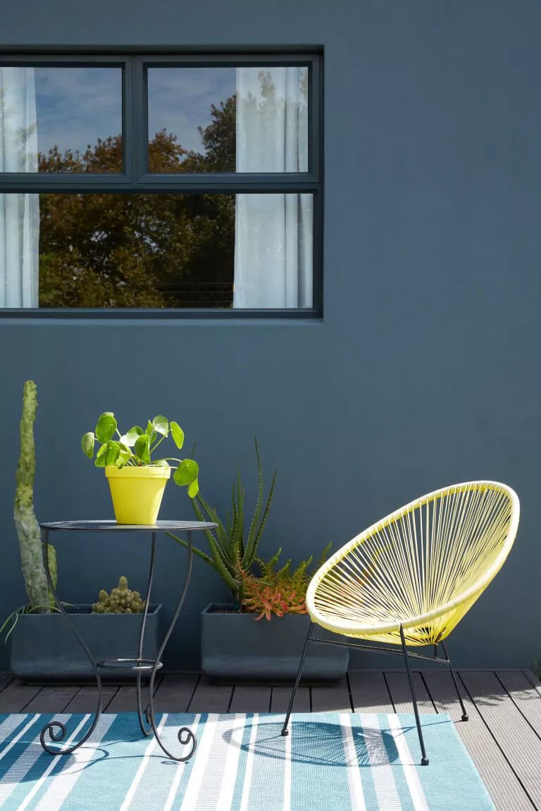 A dark grey-blue wall, with a soft blue striped outdoor rug and neon yellow chair and plant pot